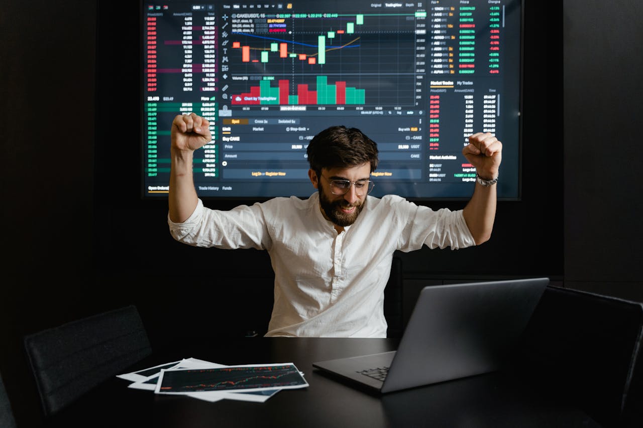 services-02 Businessman celebrates stock market success with hands raised in excitement at a trading desk.